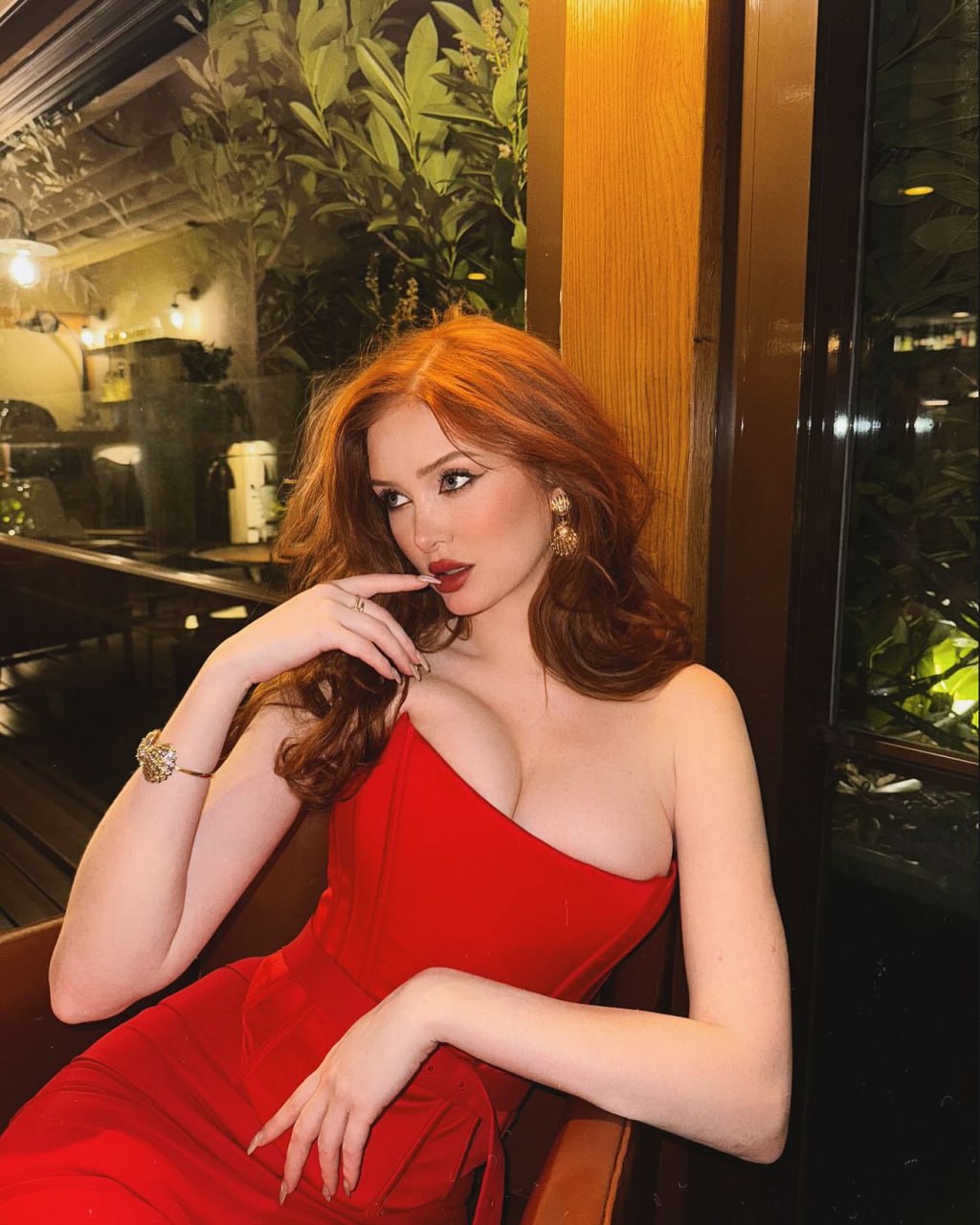 Luxury red-haired model seated elegantly in an upscale fine dining restaurant at night, wearing a sophisticated form-fitting scarlet one-shoulder evening gown with a sculpted silhouette, voluminous glossy copper-red waves styled in old Hollywood fashion, glamorous makeup with bold red lips, gold statement earrings and luxury jewelry, leaning gracefully against a polished wooden booth with a confident seductive gaze, warm ambient golden lighting, floor-to-ceiling windows reflecting the intimate restaurant interior, lush greenery outside, exclusive luxury dining atmosphere, premium cocktail lounge aesthetic, high-end fashion editorial, Vogue magazine style, cinematic composition, photorealistic skin texture, ultra-detailed fabric folds, shallow depth of field, 85mm lens, masterpiece, 8k