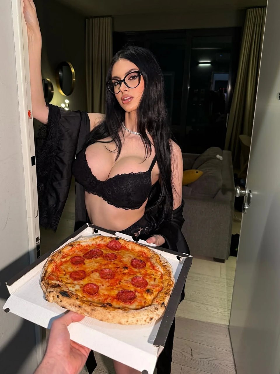Luxury brunette model answering the door of an ultra-modern five-star hotel suite at night, wearing elegant black lace lingerie beneath a silky black satin robe draped loosely off her shoulders, long straight jet-black hair, stylish oversized black eyeglasses, delicate diamond necklace, holding a freshly delivered gourmet pepperoni pizza in an open premium pizza box, photographed from the perspective of the delivery person at the doorway, warm cinematic indoor lighting, sophisticated luxury hotel interior with designer furniture, playful late-night room service atmosphere, high-end lifestyle editorial, Vogue-inspired fashion photography, photorealistic skin texture, ultra-detailed lace and satin fabrics, realistic food textures, shallow depth of field, 35mm lens, cinematic composition, masterpiece, 8k