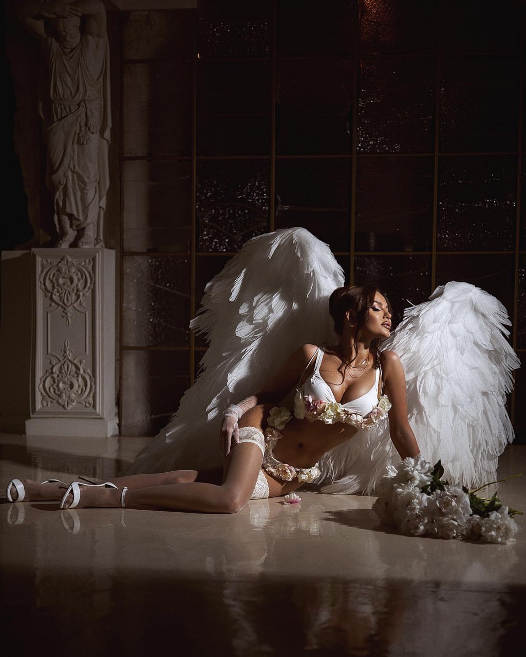 Ultra-realistic haute couture fantasy editorial of an elegant woman kneeling gracefully on a polished marble floor inside a grand neoclassical palace at night, wearing luxurious white couture lingerie with delicate floral embellishments, lace stockings, satin high heels, sheer opera gloves, and enormous handcrafted white feather angel wings spread dramatically behind her. Soft bouquet of white flowers resting beside her, classical marble statue, floor-to-ceiling dark windows with rain droplets outside, warm cinematic spotlight creating dramatic chiaroscuro lighting, reflections on polished marble floor, timeless luxury atmosphere, ethereal beauty, divine femininity, romantic fine-art fashion, Vogue Italia × Elie Saab × Zuhair Murad × Dolce & Gabbana Alta Moda campaign style, medium-format Hasselblad X2D, 80mm lens, shallow depth of field, HDR, photorealistic feathers, intricate couture detailing, realistic skin texture, elegant composition, masterpiece, ultra-photorealistic, luxury editorial, 8K