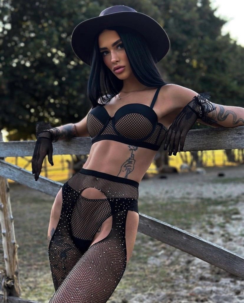 Ultra-realistic luxury western fashion editorial featuring an elegant brunette woman wearing a black fishnet lingerie set, wide-brim hat, sheer gloves, rustic wooden fence, countryside setting, golden hour lighting, cinematic composition, photorealistic, luxury lifestyle, 8k.