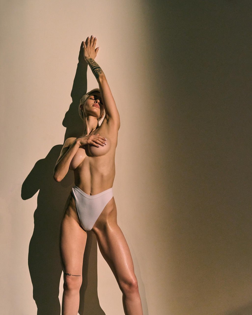 Ultra-realistic luxury fine art fashion editorial featuring an elegant blonde woman in minimalist white designer bottoms, dramatic golden spotlight, sculptural posing, clean neutral studio backdrop, artistic shadow play, premium high-fashion photography, refined luxury aesthetic.