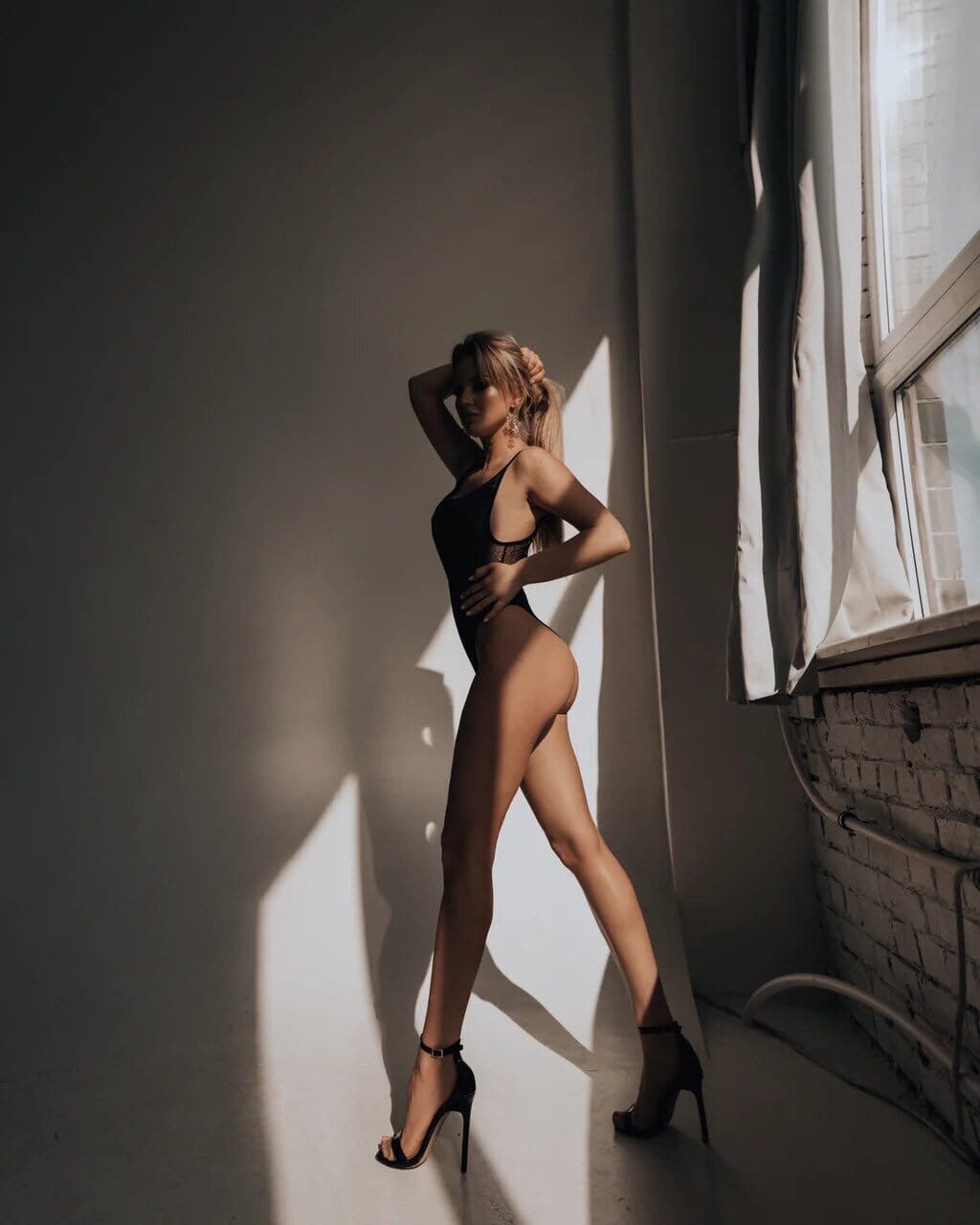 Ultra-realistic luxury fashion editorial featuring an elegant blonde woman in a black high-cut bodysuit and black stiletto heels, posing in profile inside a minimalist photo studio with dramatic natural window light, long legs, toned physique, cinematic shadows, modern editorial photography, premium luxury aesthetic, soft warm tones, high-end magazine quality, 8K.