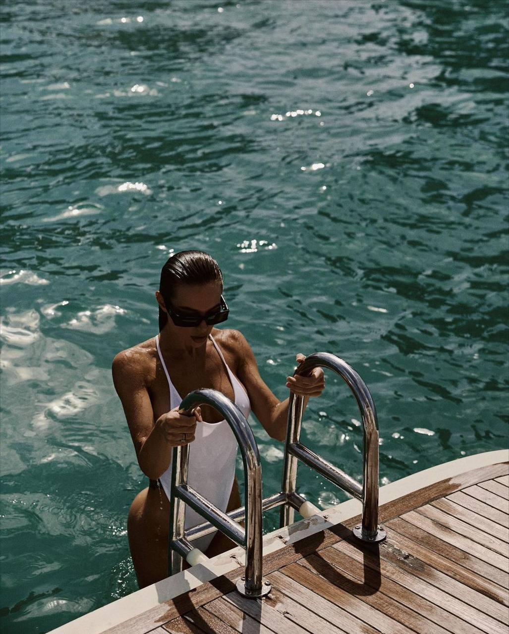 Ultra-realistic luxury yacht lifestyle editorial featuring an elegant woman emerging from crystal-clear turquoise water via a polished stainless-steel swim ladder onto a premium teak yacht deck. She wears a sleek white one-piece swimsuit and oversized black sunglasses, with wet slicked-back hair and water droplets on sun-kissed skin. Bright Mediterranean midday sun, rich teal ocean, cinematic high-end resort atmosphere, minimalist luxury aesthetic, fashion magazine editorial, natural reflections, premium travel campaign, realistic proportions, ultra-detailed textures, soft contrast, shallow depth of field, 8K photorealism.