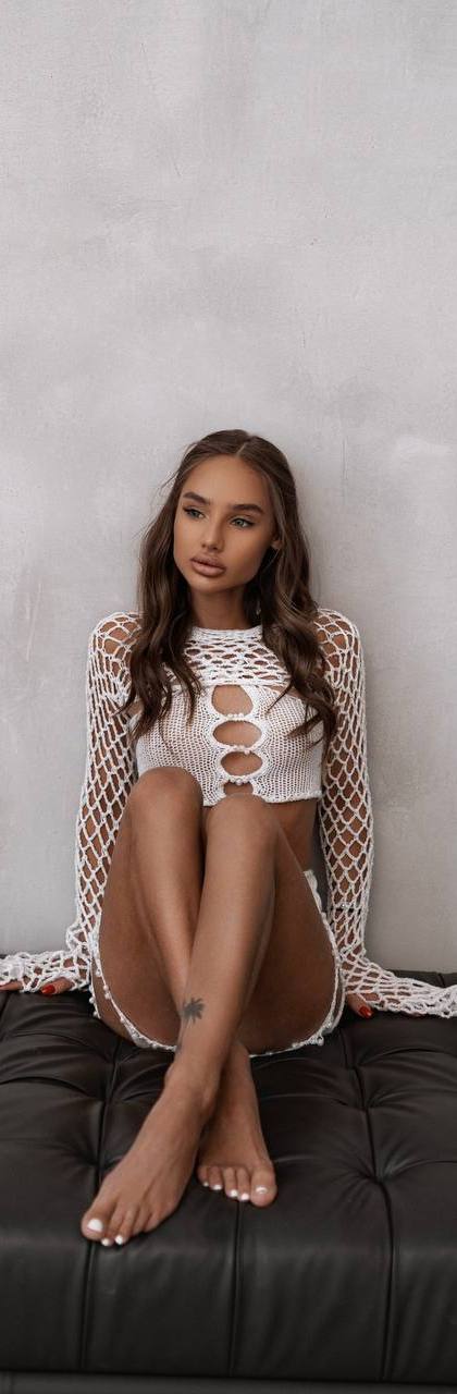 Young female model wearing a white crochet dress sitting on a black leather sofa during a fashion photoshoot.