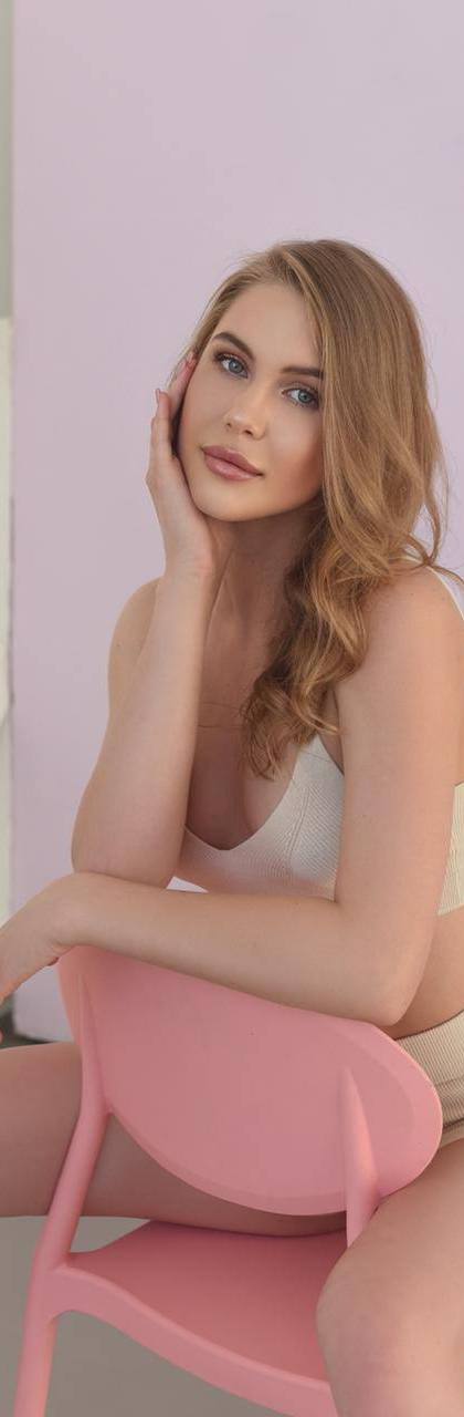 Young woman with wavy honey-blonde hair in a beige knit top and shorts sitting on a pink chair against a pink studio wall