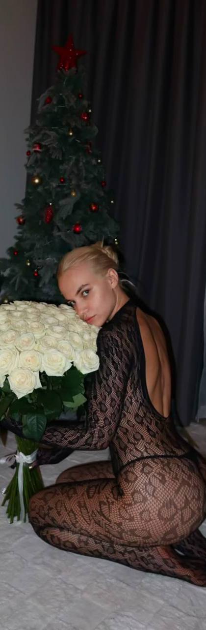 Beautiful blonde fashion model wearing a sheer black lace bodysuit with an open-back design, kneeling on a luxury bed while holding a massive bouquet of white roses, elegant modern apartment with Christmas tree in the background, sensual editorial photography, premium lifestyle aesthetic.