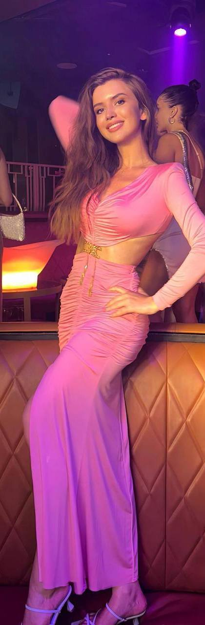 Beautiful fashion model with long wavy light brown hair wearing a luxury pink evening gown with waist cut-out, posing inside an exclusive nightclub with elegant neon purple lighting, VIP atmosphere, luxury lifestyle, high-end fashion editorial, cinematic lighting, ultra realistic, Vogue style, 85mm lens.