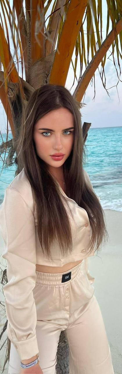 Ultra realistic portrait of a stunning young woman with long silky dark brown hair and striking blue eyes wearing a beige satin co-ord outfit while posing under a palm tree on a luxury tropical beach with turquoise water, luxury island resort, natural daylight, photorealistic, 8k.