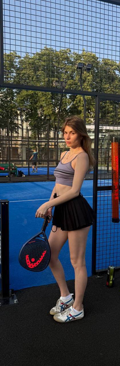 Woman in grey crop top and black pleated skirt holding a padel racket on a blue padel court