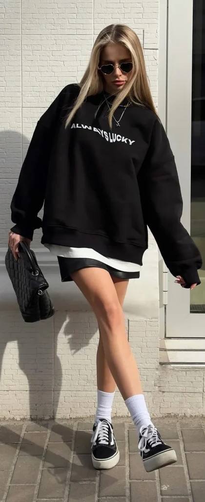 Blonde woman in oversized black sweatshirt, leather mini skirt, white socks and black sneakers posing on the street