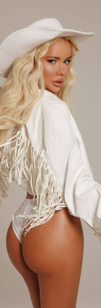 Beautiful blonde glamour model wearing a white cowboy hat and fringe jacket during a luxury studio fashion photoshoot.