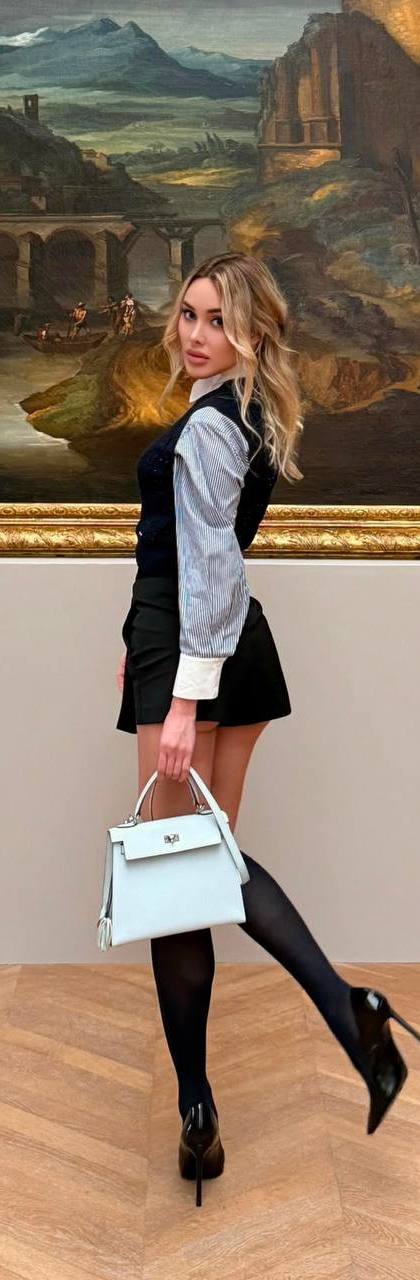 Elegant blonde fashion model wearing a navy knit top, pleated mini skirt and knee-high stockings posing inside a luxury art museum.