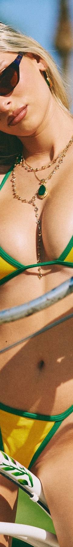 Blonde in a yellow bikini with green trim, sunglasses and layered gold necklaces posing with a vintage white bicycle among yellow flowers
