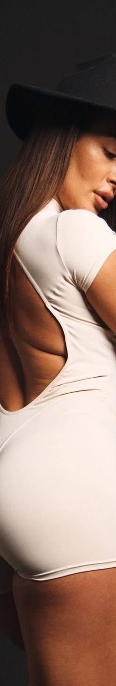 Brunette woman in a white backless bodysuit and black hat posing in a studio setting