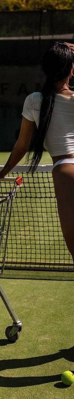 Woman standing on a tennis court beside a cart full of tennis balls and rackets, wearing athletic-inspired casual clothing, photographed от behind in bright outdoor sunlight.