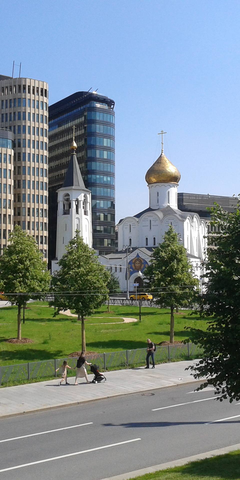 Ultra-realistic urban cityscape featuring the Cathedral of St. George on Pskov Hill surrounded by modern business architecture in central Moscow. Historic white Orthodox church with a golden dome standing in harmony among contemporary glass office buildings, landscaped green park, tree-lined boulevard, clean sidewalks, bright summer midday sunlight, deep blue cloudless sky, vibrant natural colors, realistic shadows, premium architectural photography, luxury city editorial, cinematic composition, ultra-detailed textures, HDR, photorealistic 8K, sharp focus, wide-angle perspective, modern metropolis blending history and innovation.