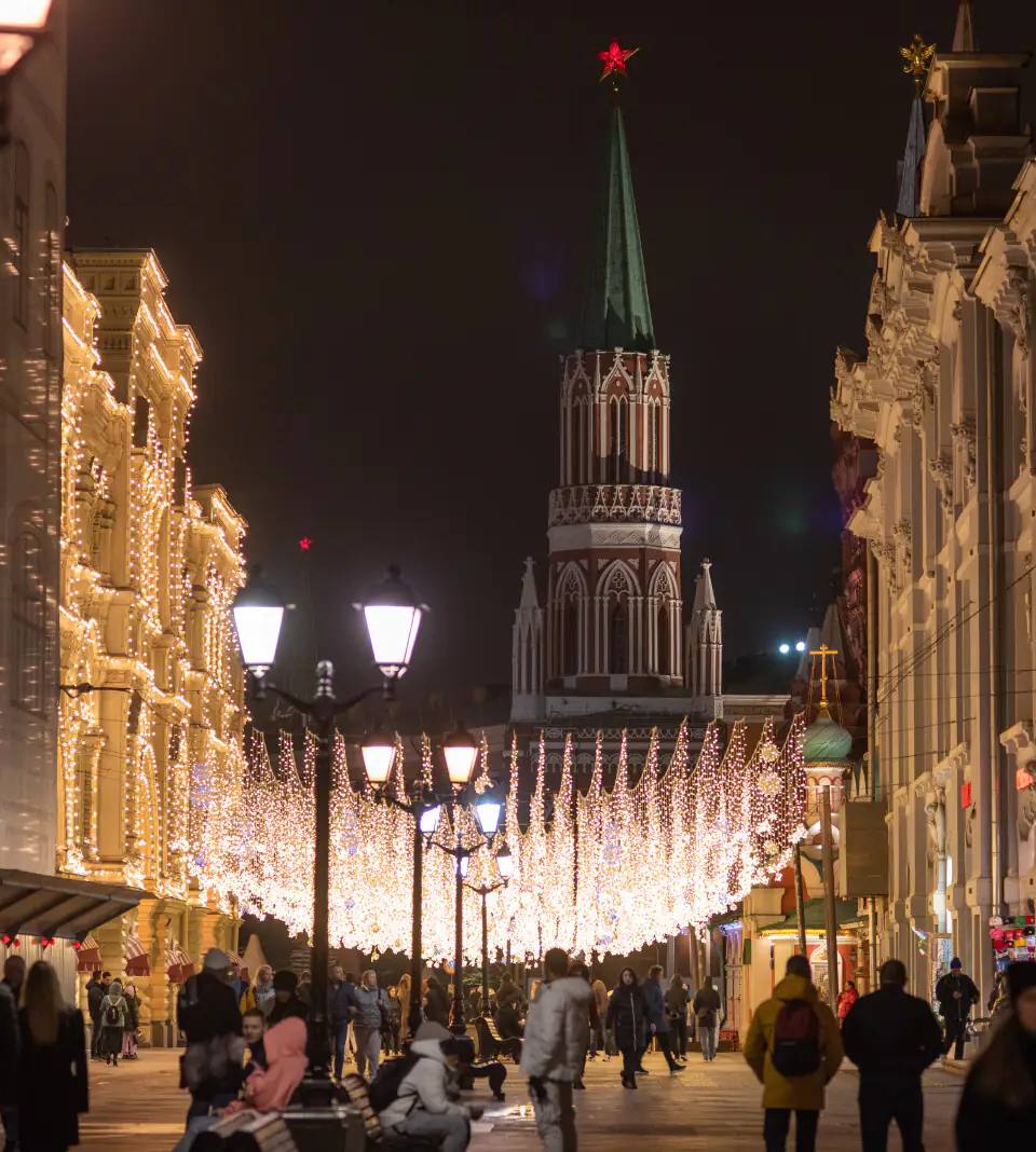 Ultra-realistic nighttime street scene on Nikolskaya Street in central Moscow during the festive season. Elegant historic European architecture illuminated with thousands of warm white decorative lights suspended across the pedestrian boulevard. The iconic Kremlin tower with its glowing red star rising dramatically in the background beneath a clear night sky. Stylish pedestrians strolling through the lively street, vintage black street lamps casting warm ambient light, luxury holiday atmosphere, cinematic symmetry, premium travel editorial photography, photorealistic 8K, HDR, ultra-detailed textures, realistic reflections, rich architectural details, festive urban elegance, high-end cityscape composition.