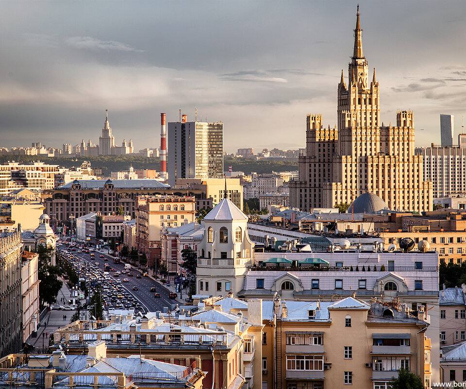Ultra-realistic panoramic cityscape of central Moscow at golden hour viewed from an elevated rooftop perspective. The iconic Ministry of Foreign Affairs skyscraper (one of the Seven Sisters) dominates the skyline, surrounded by elegant historic buildings, wide boulevards filled with traffic, classical European architecture blending with modern skyscrapers in the distance. Warm sunset light casting long shadows across the city, dramatic cloudy sky with soft golden highlights, premium urban atmosphere, cinematic composition, luxury travel editorial photography, photorealistic 8K, HDR, ultra-detailed architecture, realistic lighting, crisp textures, vibrant natural colors, high-end cityscape, timeless metropolitan elegance.