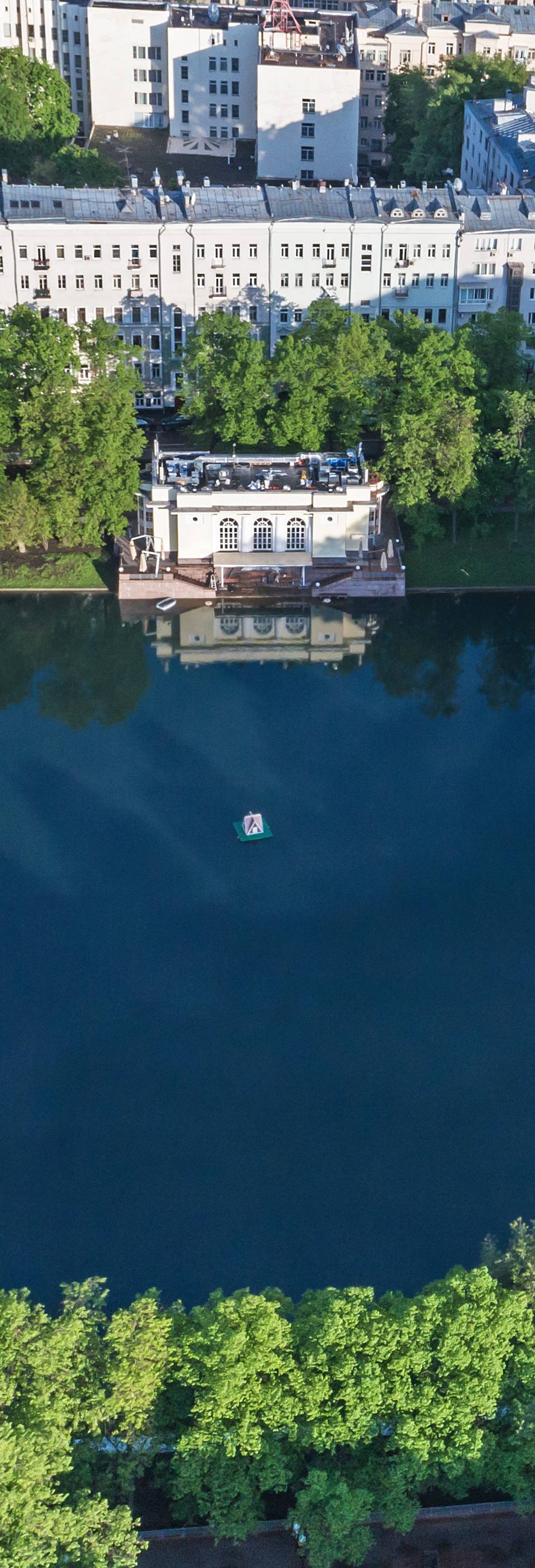Ultra-realistic aerial drone view of Patriarch Ponds in central Moscow during a bright summer morning. A perfectly rectangular historic pond surrounded by lush green trees, elegant walking paths, and prestigious residential buildings with classic European architecture. Calm mirror-like water reflecting the surrounding landscape, vibrant foliage, peaceful urban oasis in the heart of the city, luxury residential atmosphere, premium aerial photography, cinematic top-down composition, photorealistic 8K, HDR, ultra-detailed textures, crisp natural lighting, realistic colors, high-end travel editorial, sophisticated metropolitan park, exclusive neighborhood.