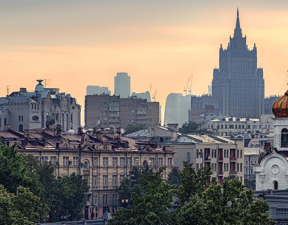 Ultra-realistic panoramic cityscape of historic central Moscow at sunrise, blending elegant classical architecture with iconic Soviet-era landmarks. The majestic Ministry of Foreign Affairs skyscraper (one of the Seven Sisters) rises dramatically above the city skyline, while historic Orthodox church domes with golden copper roofs and ornate European buildings fill the foreground. Soft golden morning light, gentle atmospheric haze, layered urban skyline fading into the distance, lush green treetops framing the lower city, cinematic depth, timeless metropolitan elegance, luxury travel editorial photography, photorealistic 8K, HDR, ultra-detailed architectural textures, realistic natural lighting, premium magazine quality, sophisticated European city atmosphere, wide panoramic composition.