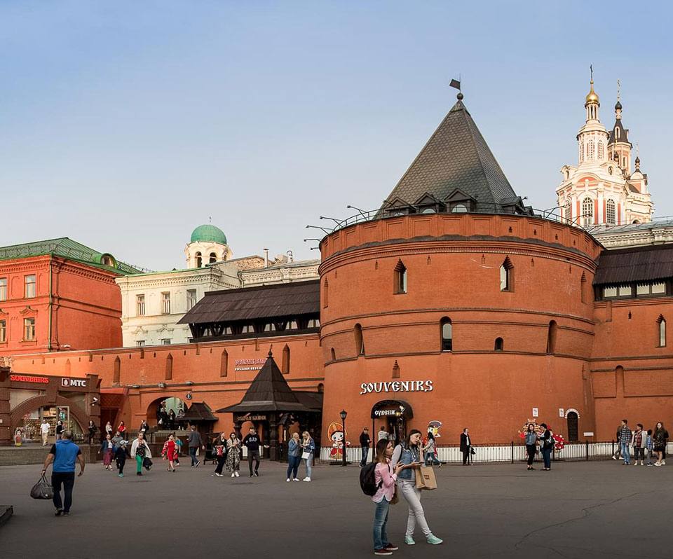 Ultra-realistic architectural street scene of the historic Kitay-Gorod fortress walls in central Moscow during warm golden hour. Massive circular red-brick medieval tower with a dark conical roof standing prominently in a spacious pedestrian square, surrounded by beautifully restored historic buildings with colorful facades and elegant Orthodox church towers rising in the background. Visitors strolling through the square, lively yet refined atmosphere, soft golden sunlight illuminating the brick textures, clear blue sky, timeless Russian architectural heritage blended with modern city life, cinematic composition, premium travel editorial photography, photorealistic 8K, HDR, ultra-detailed masonry, realistic lighting, crisp architectural textures, luxury cultural destination, sophisticated urban landmark.