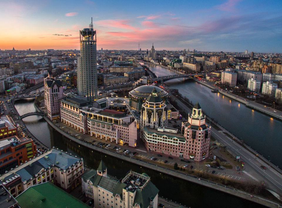 Ultra-realistic aerial view of Moscow at blue hour, overlooking the prestigious Balchug district and the Moscow River. Elegant waterfront lined with luxurious residential and commercial architecture, iconic modern high-rise tower dominating the skyline, beautifully illuminated bridges crossing the river, reflections shimmering across calm water, sophisticated urban atmosphere, warm city lights blending with the cool twilight sky painted in soft pink and deep blue tones. Dense metropolitan skyline stretching to the horizon, premium financial district, cinematic composition, luxury travel editorial photography, photorealistic 8K, HDR, ultra-detailed buildings, realistic reflections, atmospheric evening lighting, breathtaking cityscape, high-end architectural photography.