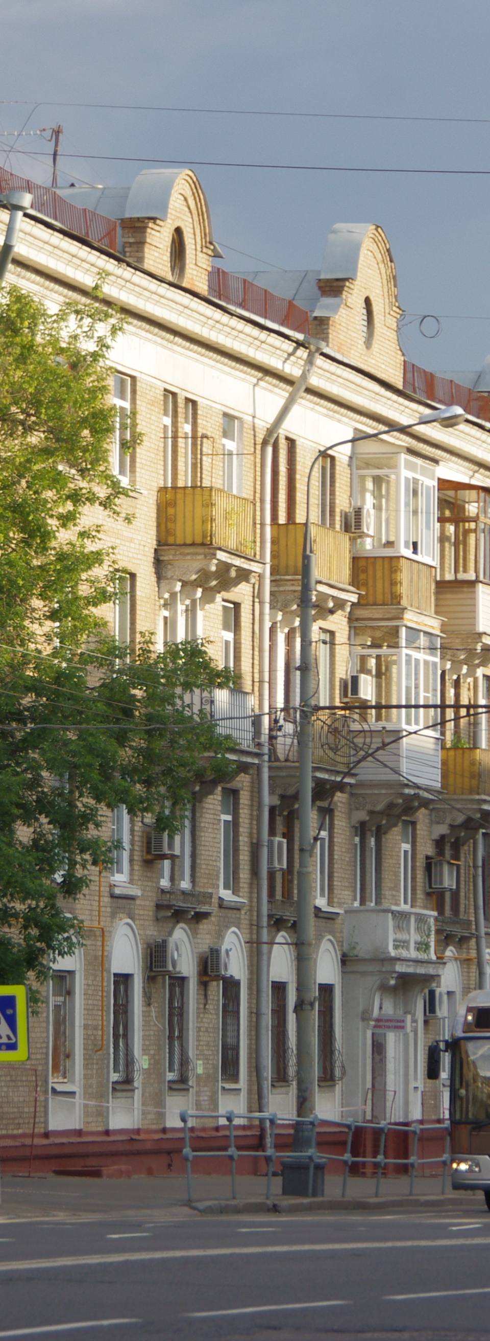 Ultra-realistic street view of a prestigious residential district in central Moscow during golden hour. Wide boulevard with elegant Stalin-era apartment buildings featuring cream-colored brick facades, classic balconies and decorative rooflines, lush mature trees lining both sides of the avenue, modern electric trolleybus traveling along a nearly empty road with overhead power lines, warm evening sunlight illuminating the architecture while dramatic storm clouds gather in the distance. Clean sidewalks, refined urban atmosphere, premium European city aesthetic, cinematic composition, luxury editorial travel photography, photorealistic 8K, HDR, ultra-detailed architecture, realistic lighting, natural colors, atmospheric depth, high-end architectural photography.