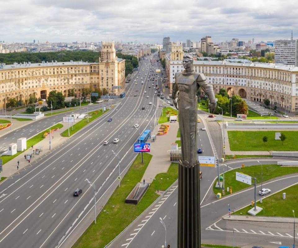 Ultra-realistic aerial view of one of Moscow's grand central avenues featuring a monumental boulevard lined with iconic Stalinist architecture. Massive cream-colored residential buildings symmetrically framing a wide multi-lane avenue stretching toward the horizon, elegant green medians and landscaped public spaces, imposing bronze monument rising above the cityscape, light traffic creating a calm metropolitan atmosphere. Dramatic overcast sky with soft diffused daylight highlighting the scale of the architecture, panoramic skyline of central Moscow in the distance. Luxury urban editorial photography, photorealistic 8K, HDR, ultra-detailed buildings, realistic textures, cinematic composition, premium architectural photography, atmospheric depth, natural colors, breathtaking cityscape.