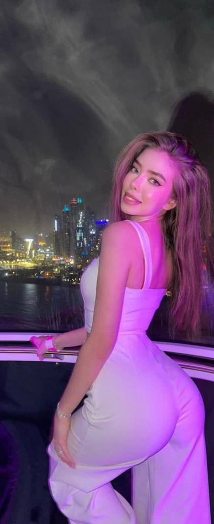 Woman with long hair in a white jumpsuit looking over her shoulder inside a glass observation wheel cabin with night city lights behind