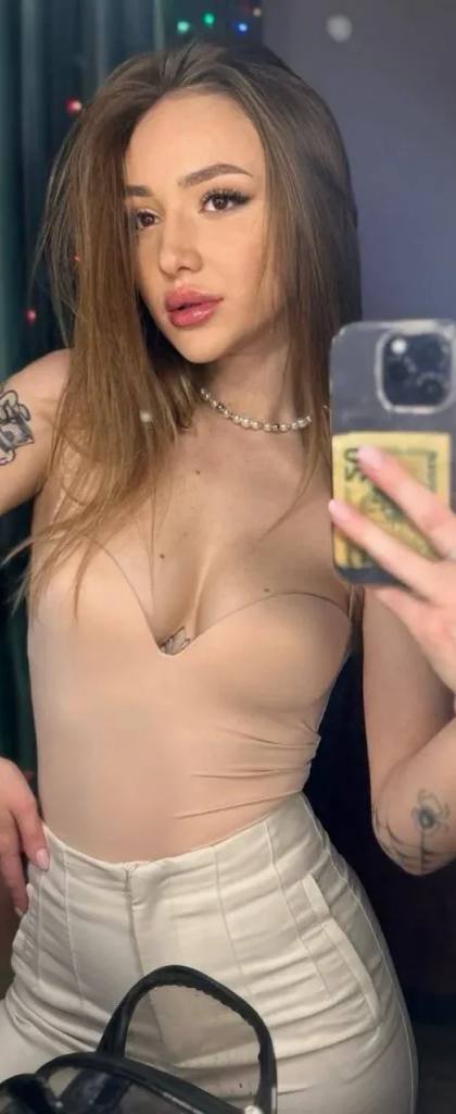 Mirror selfie of a tattooed woman in a beige strapless bodysuit, white trousers and a pearl necklace