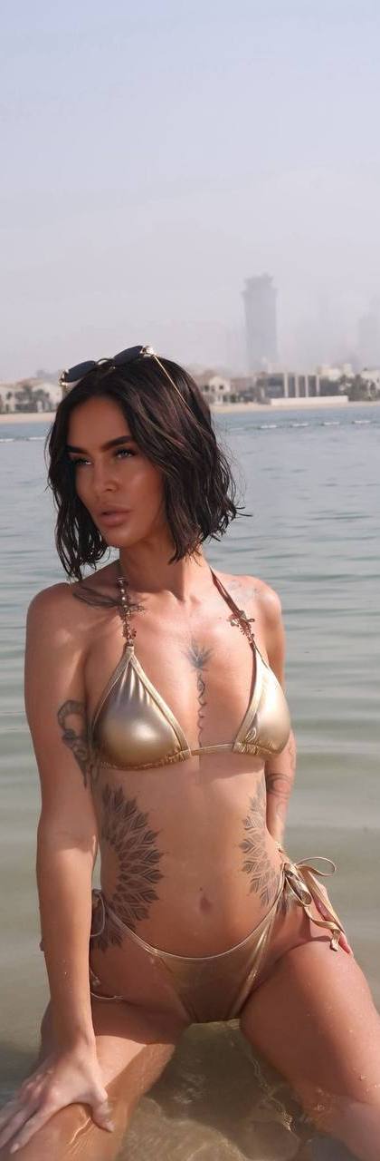 Ultra realistic portrait of a glamorous tattooed brunette woman with a short black bob haircut wearing a metallic gold bikini while posing in shallow crystal clear water on a luxury beach in Moscow, soft natural sunlight, luxury travel photography, photorealistic, 8k.