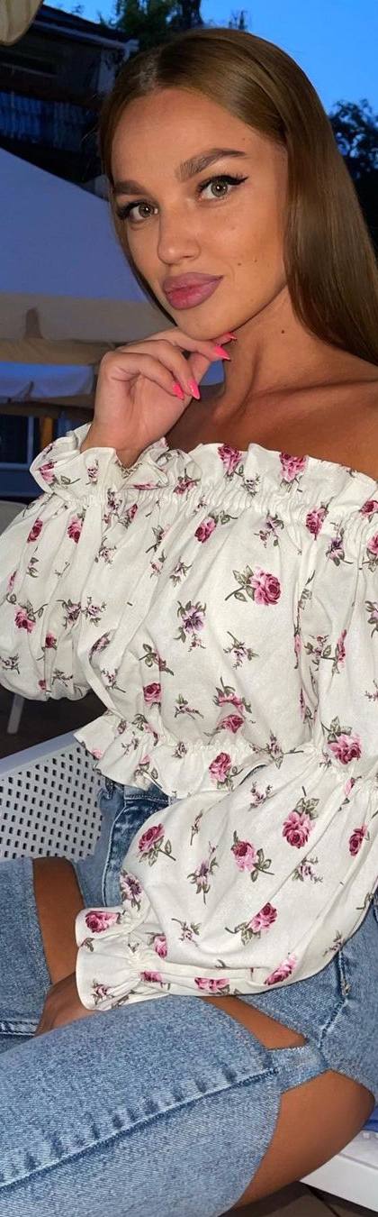 Blonde woman in a white off-shoulder floral blouse and ripped jeans sitting in a chair by an outdoor pool in the evening